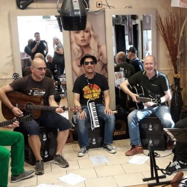 5 Musiker spielen sitzend im Friseursalon und im Spiegel dahinter sieht man etwas Publikum